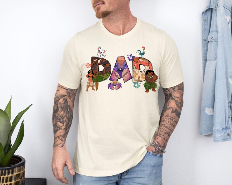 K&ouml;nnte beinhalten: Ein cremefarbenes T-Shirt mit einem Aufdruck des Wortes "DAD", das aus Figuren aus dem Film Vaiana besteht. Die Figuren sind Maui, Vaiana, das Schwein Pua, der Hahn Hei Hei und der Fischhaken des Halbgottes Maui.