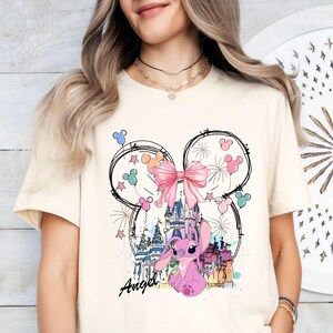 May include: Cream-colored t-shirt featuring a whimsical design. The graphic includes a pink cartoon character, a castle, fireworks, and a large pink bow. The word "Angel" is written in cursive below the character. The design is set within a Mickey Mouse ear outline.