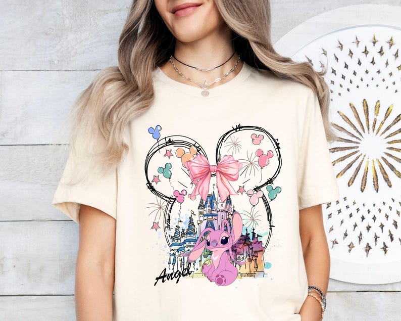 May include: Cream-colored t-shirt featuring a whimsical design. The graphic includes a pink cartoon character, a castle, fireworks, and a large pink bow. The word "Angel" is written in cursive below the character. The design is set within a Mickey Mouse ear outline.