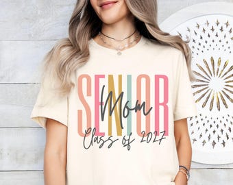 Op de afbeelding: Een crèmekleurig T-shirt met korte mouwen met het woord SENIOR in grote, verticale, veelkleurige blokletters in tinten perzik, geel, blauwgroen en roze. Het woord Mom en de tekst Class of 2027 zijn gedrukt in een donkergrijs cursief lettertype.