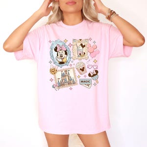 Pu&ograve; includere: T-shirt rosa chiaro con un design a collage di cartoni animati. Il design include Minnie Mouse, un castello, popcorn, gelato e la parola "MAGIC". Le illustrazioni sono incorniciate con bordi decorativi e stelle.