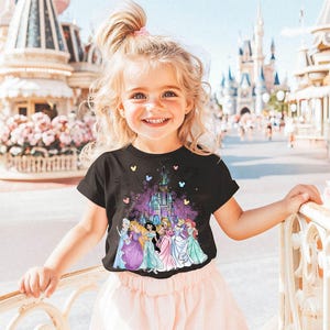Puede incluir: Una ni&ntilde;a peque&ntilde;a con una camiseta negra con un gr&aacute;fico colorido de princesas Disney y el castillo de Cenicienta. La camiseta tiene un esquema de color rosa, morado y azul. La ni&ntilde;a est&aacute; de pie frente a una barandilla blanca con un arbusto de flores rosas en el fondo.