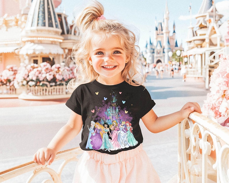 Puede incluir: Una ni&ntilde;a peque&ntilde;a con una camiseta negra con un gr&aacute;fico colorido de princesas Disney y el castillo de Cenicienta. La camiseta tiene un esquema de color rosa, morado y azul. La ni&ntilde;a est&aacute; de pie frente a una barandilla blanca con un arbusto de flores rosas en el fondo.