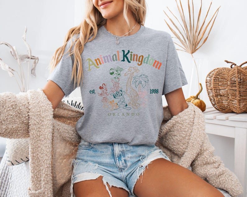 K&ouml;nnte beinhalten: Ein meliertes graues T-Shirt mit dem Text "Animal Kingdom" in bunten Buchstaben, Cartoon-Tierfiguren und dem Text "est. 1998 ORLANDO". Das T-Shirt hat kurze &Auml;rmel.