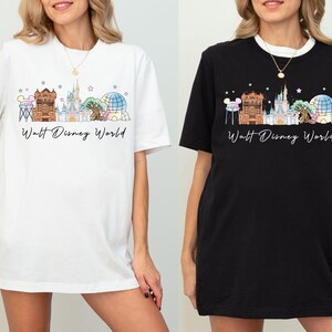 May include: A white t-shirt and a black t-shirt, each featuring a graphic print of the Walt Disney World skyline. The design includes iconic park landmarks in pastel colors, small stars, and the text Walt Disney World in a black cursive script.
