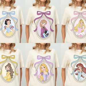 May include: Six cream-colored t-shirts with a different Disney princess in each. Each princess is in a pearl-like oval frame with a pink, purple, blue, or yellow bow at the top. The princesses are Snow White, Elsa, Aurora, Belle, Rapunzel, and Ariel.