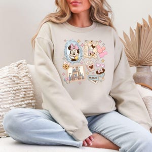 Pu&ograve; includere: Felpa beige con un colorato collage di immagini di cartoni animati. Il design include Minnie Mouse, popcorn, un castello e la parola "MAGIC". La felpa ha maniche lunghe e girocollo.
