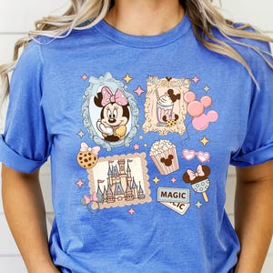 Pu&ograve; includere: T-shirt blu con una grafica colorata raffigurante Minnie Mouse, un castello, popcorn e gelato. Il design include la parola "MAGIC" e altri elementi a tema, il tutto incorniciato da stelle e bordi decorativi.