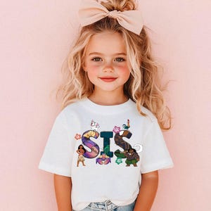 K&ouml;nnte beinhalten: Ein wei&szlig;es T-Shirt mit einer bunten Grafik des Wortes "SIS", die Figuren aus dem Animationsfilm Vaiana zeigt. Die Grafik enth&auml;lt Vaiana, Maui, Pua das Schwein und Hei Hei den Hahn.