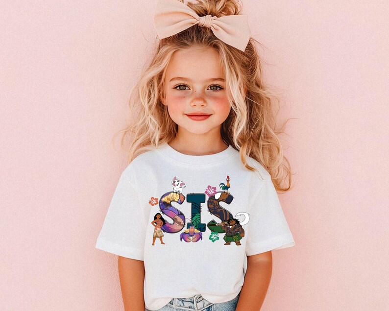 K&ouml;nnte beinhalten: Ein wei&szlig;es T-Shirt mit einer bunten Grafik des Wortes "SIS", die Figuren aus dem Animationsfilm Vaiana zeigt. Die Grafik enth&auml;lt Vaiana, Maui, Pua das Schwein und Hei Hei den Hahn.