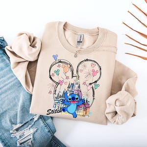 May include: A beige sweatshirt featuring a graphic of Stitch from Lilo & Stitch, with a castle, fireworks, and Mickey Mouse ears design. The word "Stitch" is written in black script. The sweatshirt is paired with blue jeans.