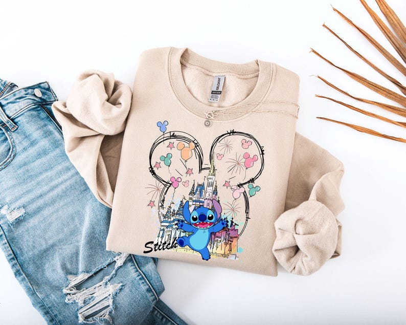 May include: A beige sweatshirt featuring a graphic of Stitch from Lilo & Stitch, with a castle, fireworks, and Mickey Mouse ears design. The word "Stitch" is written in black script. The sweatshirt is paired with blue jeans.