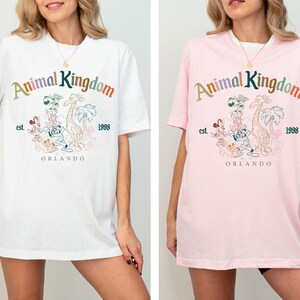 K&ouml;nnte beinhalten: Wei&szlig;e und rosa T-Shirts mit dem Schriftzug "Animal Kingdom" in bunten Buchstaben, zusammen mit einer Grafik verschiedener Zeichentrickfiguren, einer Giraffe und Palmen. Die Shirts enthalten auch den Text "est. 1998" und "Orlando".