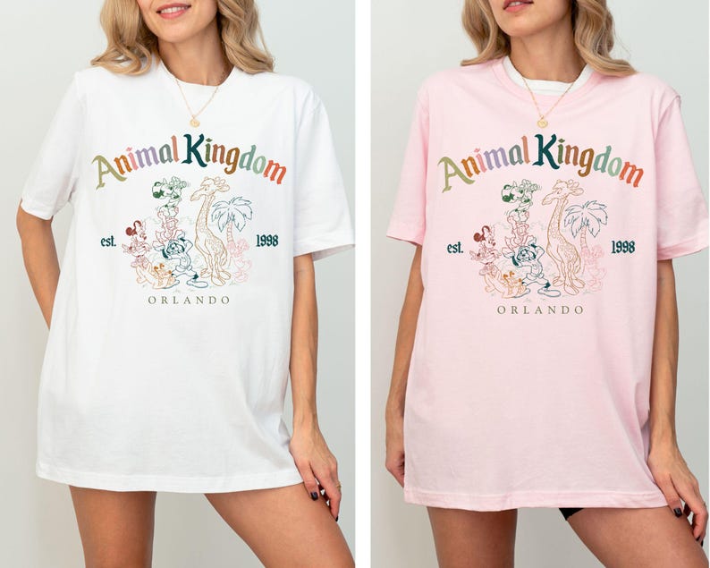 K&ouml;nnte beinhalten: Wei&szlig;e und rosa T-Shirts mit dem Schriftzug "Animal Kingdom" in bunten Buchstaben, zusammen mit einer Grafik verschiedener Zeichentrickfiguren, einer Giraffe und Palmen. Die Shirts enthalten auch den Text "est. 1998" und "Orlando".