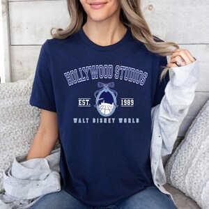 Puede incluir: Camiseta azul marino con el texto "HOLLYWOOD STUDIOS" en letras blancas arqueadas. Debajo, un gr&aacute;fico de una silueta de edificio y un globo, con el texto "EST. 1989 WALT DISNEY WORLD" en blanco.