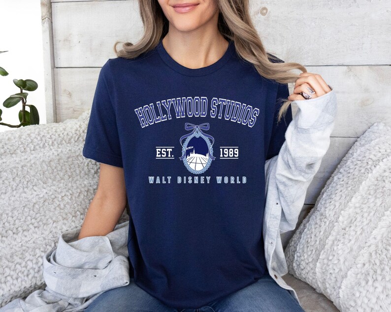 Puede incluir: Camiseta azul marino con el texto "HOLLYWOOD STUDIOS" en letras blancas arqueadas. Debajo, un gr&aacute;fico de una silueta de edificio y un globo, con el texto "EST. 1989 WALT DISNEY WORLD" en blanco.