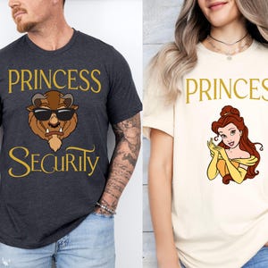 以下が含まれることがあります： 2枚のTシャツ。1枚は濃いグレーで、「PRINCESS SECURITY」とサングラスをかけた漫画の獣が描かれています。もう1枚はクリーム色で、「PRINCESS」と漫画のプリンセスが描かれています。どちらのテキストも金色です。