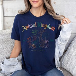 K&ouml;nnte beinhalten: Marineblaues T-Shirt mit der Aufschrift "Animal Kingdom" in bunten Buchstaben. Das Shirt zeigt eine Grafik einer Giraffe und anderer Tiere, mit "Orlando" und "1998" darunter gedruckt.