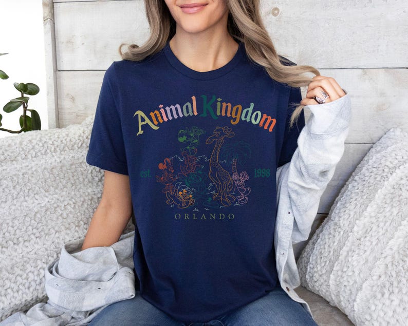 K&ouml;nnte beinhalten: Marineblaues T-Shirt mit der Aufschrift "Animal Kingdom" in bunten Buchstaben. Das Shirt zeigt eine Grafik einer Giraffe und anderer Tiere, mit "Orlando" und "1998" darunter gedruckt.