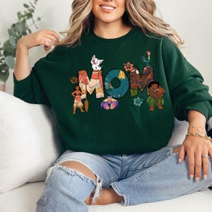 K&ouml;nnte beinhalten: Gr&uuml;ner Pullover mit einem Grafikdruck von Figuren aus dem Disney-Film Vaiana. Die Grafik enth&auml;lt das Wort "MOM" mit den Figuren, die die Buchstaben umgeben.