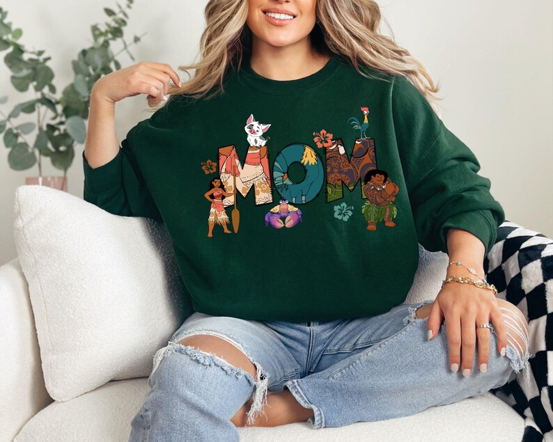 K&ouml;nnte beinhalten: Gr&uuml;ner Pullover mit einem Grafikdruck von Figuren aus dem Disney-Film Vaiana. Die Grafik enth&auml;lt das Wort "MOM" mit den Figuren, die die Buchstaben umgeben.