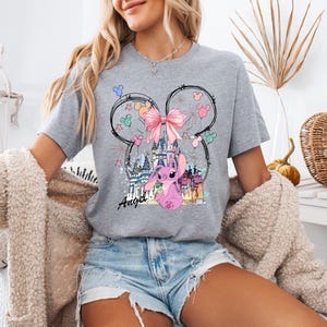 May include: A gray t-shirt featuring a graphic of a pink cartoon character, a castle, and a pink bow. The design is set within a Mickey Mouse head outline. The word "Angel" is written in cursive below the character.