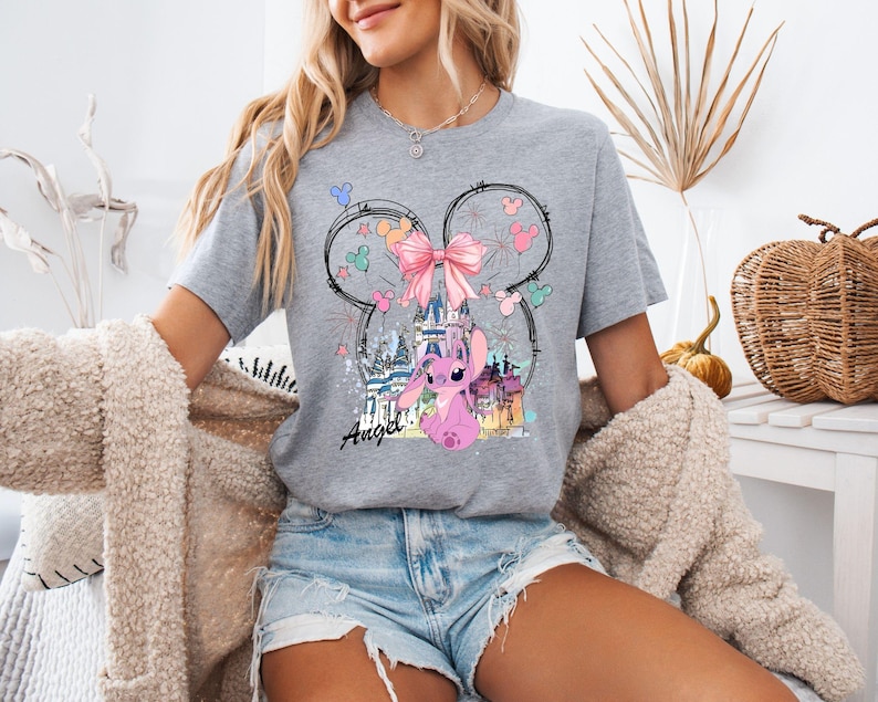 May include: A gray t-shirt featuring a graphic of a pink cartoon character, a castle, and a pink bow. The design is set within a Mickey Mouse head outline. The word "Angel" is written in cursive below the character.