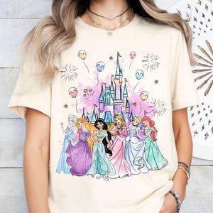 Puede incluir: Una camiseta de color crema con un gr&aacute;fico de las princesas de Disney frente al castillo de Cenicienta. Las princesas llevan sus vestidos caracter&iacute;sticos y hay fuegos artificiales y globos en el fondo.