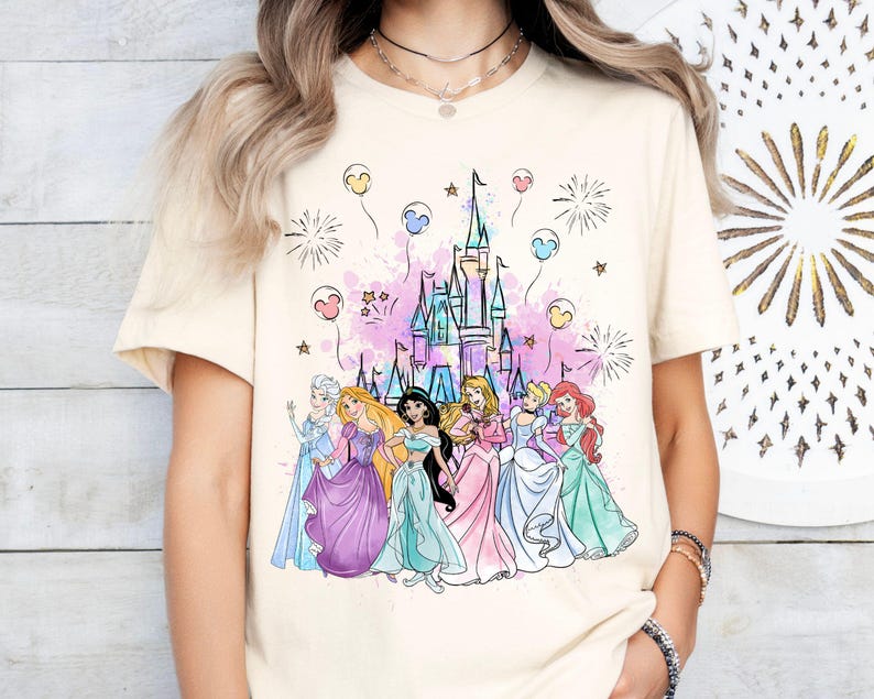 Puede incluir: Una camiseta de color crema con un gr&aacute;fico de las princesas de Disney frente al castillo de Cenicienta. Las princesas llevan sus vestidos caracter&iacute;sticos y hay fuegos artificiales y globos en el fondo.