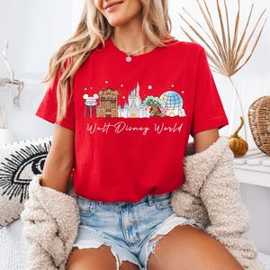 May include: A bright red short-sleeve t-shirt featuring a graphic print of the Walt Disney World skyline, including the water tower, castle, and iconic park structures, with the text Walt Disney World written in a white cursive script below.