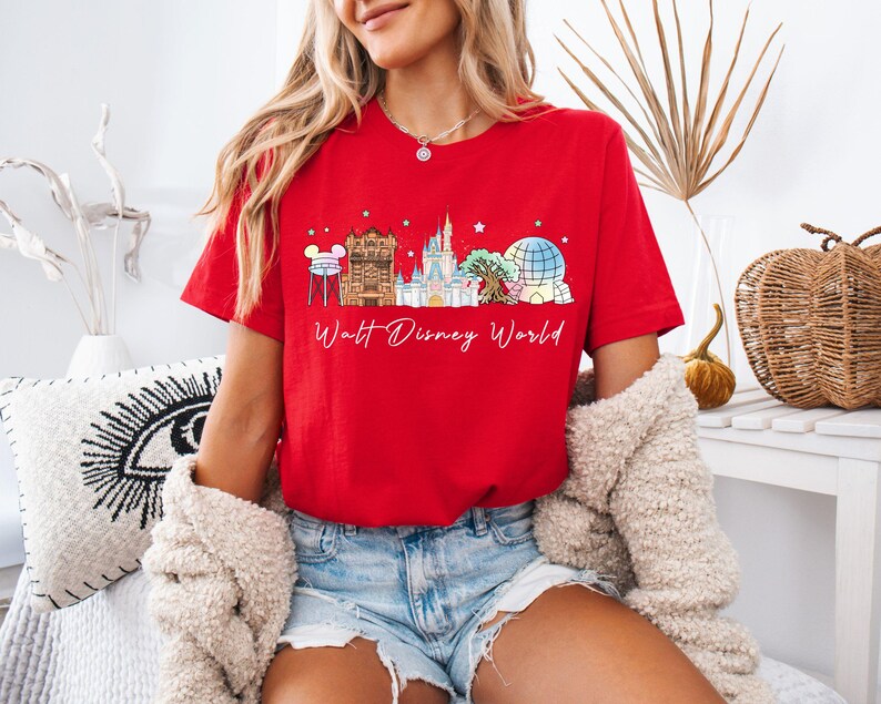 May include: A bright red short-sleeve t-shirt featuring a graphic print of the Walt Disney World skyline, including the water tower, castle, and iconic park structures, with the text Walt Disney World written in a white cursive script below.