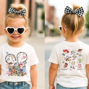 Op de afbeelding: Een wit t-shirt met een kleurrijke afbeelding van Disney-personages, waaronder Woody en Buzz Lightyear. De voorkant heeft een Mickey Mouse-hoofdontwerp met vuurwerk en ballonnen. De achterkant van het shirt toont meer karakterillustraties en tekst.