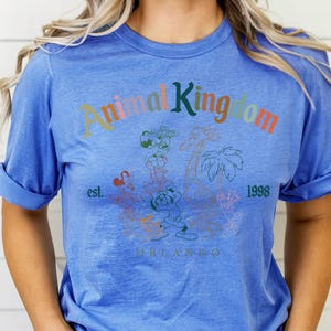 K&ouml;nnte beinhalten: Blaues T-Shirt mit hochgekrempelten &Auml;rmeln. Auf der Vorderseite steht der Text "Animal Kingdom" in verschiedenen Farben, mit einer Grafik von Zeichentieren und Palmen. Der Text "est. 1998 Orlando" ist ebenfalls zu sehen.