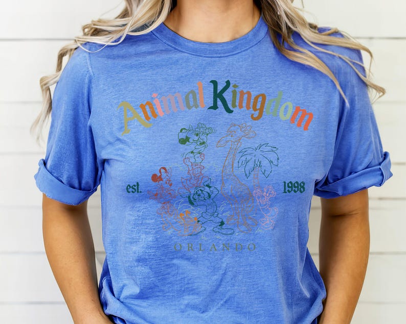 K&ouml;nnte beinhalten: Blaues T-Shirt mit hochgekrempelten &Auml;rmeln. Auf der Vorderseite steht der Text "Animal Kingdom" in verschiedenen Farben, mit einer Grafik von Zeichentieren und Palmen. Der Text "est. 1998 Orlando" ist ebenfalls zu sehen.