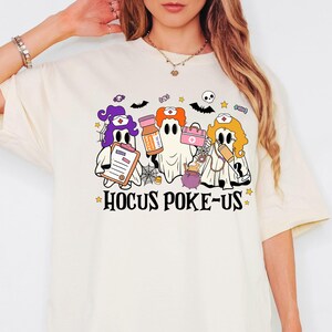 May include: Cream-colored t-shirt featuring a Halloween-themed graphic. The design includes three cartoon ghosts dressed as nurses, holding medical supplies. The text "Hocus Poke-Us" is printed below the ghosts in a stylized font, with stars.