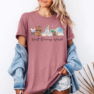 May include: A mauve short-sleeved t-shirt featuring a graphic print of various theme park landmarks and the text Walt Disney World in black script. The design includes a water tower, a theater, a castle, a tree, and a geodesic sphere.