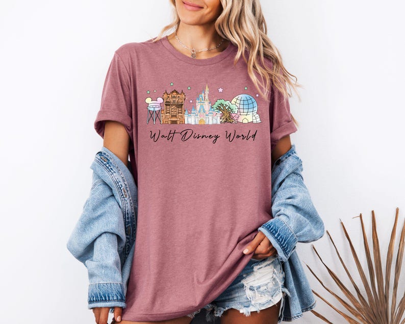 May include: A mauve short-sleeved t-shirt featuring a graphic print of various theme park landmarks and the text Walt Disney World in black script. The design includes a water tower, a theater, a castle, a tree, and a geodesic sphere.