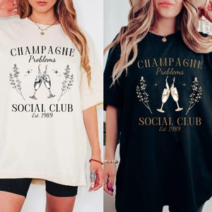 May include: Two oversized t-shirts with a graphic design featuring a champagne toast and the text "Champagne Problems Social Club Est. 1989". One shirt is white and the other is black.