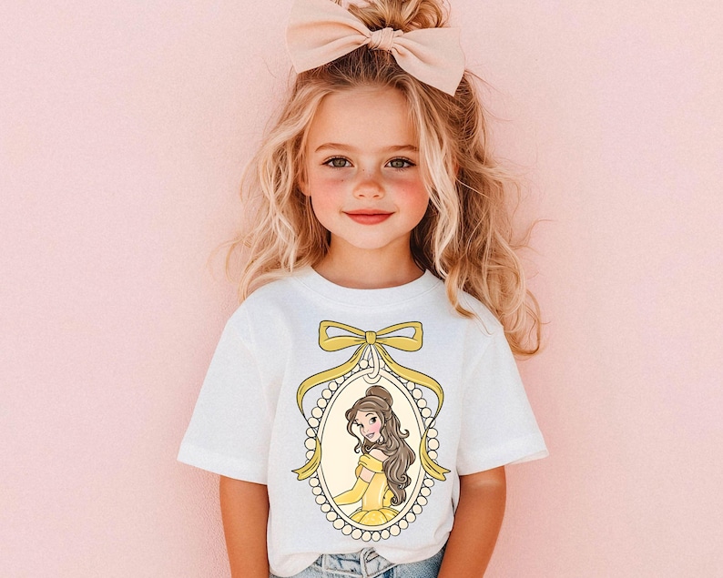 May include: A white t-shirt with a yellow and white illustration of Belle from Beauty and the Beast. The illustration is framed with a pearl necklace and a yellow bow.