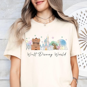 May include: A cream-colored short-sleeve t-shirt featuring a graphic print of the Walt Disney World skyline, including the water tower, Tower of Terror, Cinderella Castle, Tree of Life, and Spaceship Earth, with the text Walt Disney World below.