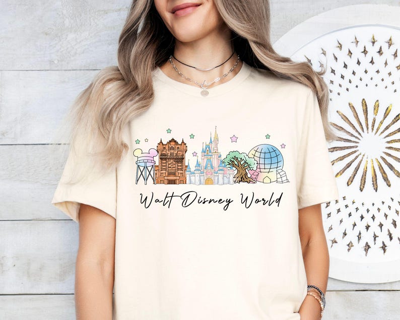 May include: A cream-colored short-sleeve t-shirt featuring a graphic print of the Walt Disney World skyline, including the water tower, Tower of Terror, Cinderella Castle, Tree of Life, and Spaceship Earth, with the text Walt Disney World below.