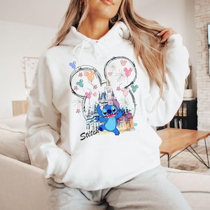 May include: White hooded sweatshirt featuring a graphic of Stitch from Lilo & Stitch, with a castle and fireworks design. The word "Stitch" is written in black script below the character. The design is framed by a Mickey Mouse ear outline.