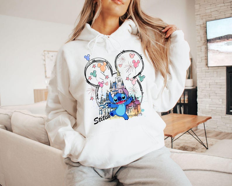 May include: White hooded sweatshirt featuring a graphic of Stitch from Lilo & Stitch, with a castle and fireworks design. The word "Stitch" is written in black script below the character. The design is framed by a Mickey Mouse ear outline.