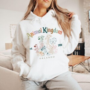 K&ouml;nnte beinhalten: Wei&szlig;er Kapuzenpullover mit dem Text "Animal Kingdom" in Regenbogenfarben, zusammen mit einer Grafik einer Giraffe, Palmen und anderen Figuren. Der Pullover hat auch den Text "est. 1998 ORLANDO".