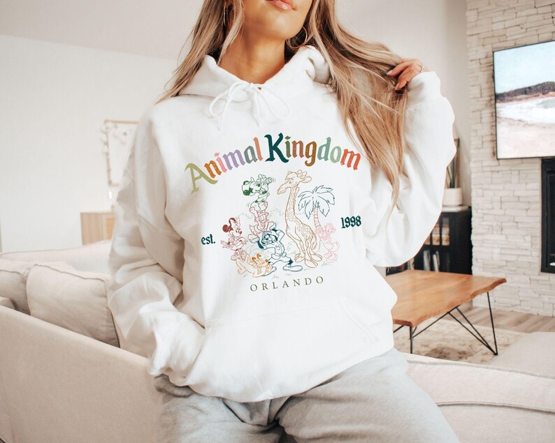 K&ouml;nnte beinhalten: Wei&szlig;er Kapuzenpullover mit dem Text "Animal Kingdom" in Regenbogenfarben, zusammen mit einer Grafik einer Giraffe, Palmen und anderen Figuren. Der Pullover hat auch den Text "est. 1998 ORLANDO".