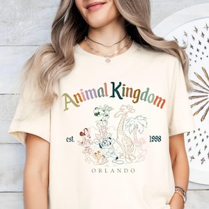K&ouml;nnte beinhalten: Cremefarbenes T-Shirt mit dem Schriftzug "Animal Kingdom" in Regenbogenfarben und Cartoon-Tierillustrationen. Der Text "est. 1998 ORLANDO" ist ebenfalls auf dem Shirt aufgedruckt. Ein l&auml;ssiges Kleidungsst&uuml;ck.