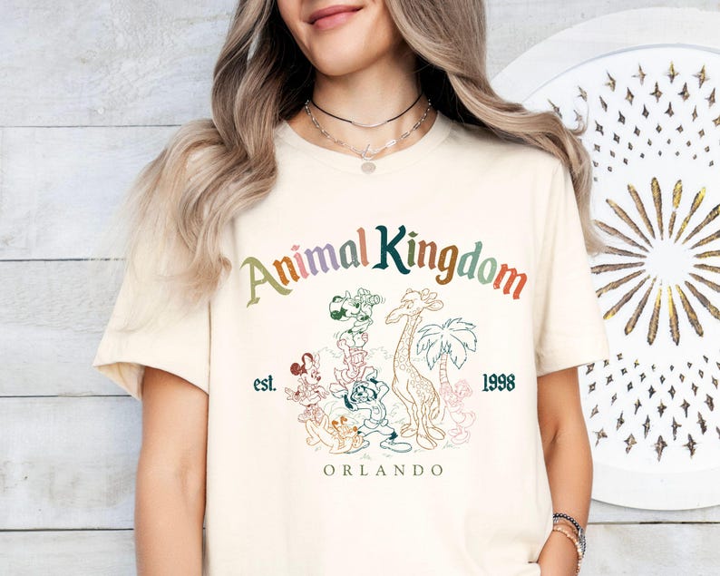K&ouml;nnte beinhalten: Cremefarbenes T-Shirt mit dem Schriftzug "Animal Kingdom" in Regenbogenfarben und Cartoon-Tierillustrationen. Der Text "est. 1998 ORLANDO" ist ebenfalls auf dem Shirt aufgedruckt. Ein l&auml;ssiges Kleidungsst&uuml;ck.