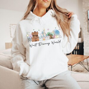 May include: A white hooded sweatshirt featuring a colorful graphic of the Walt Disney World skyline, including iconic park landmarks and the text Walt Disney World in black script. The hoodie has long sleeves, a front pocket, and a drawstring hood.