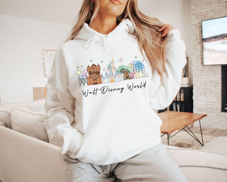 May include: A white hooded sweatshirt featuring a colorful graphic of the Walt Disney World skyline, including iconic park landmarks and the text Walt Disney World in black script. The hoodie has long sleeves, a front pocket, and a drawstring hood.