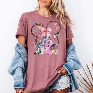 May include: A mauve t-shirt featuring a graphic of a pink cartoon character, a castle, and a pink bow. The design is framed by a Mickey Mouse ear outline with colorful accents. The word "Angel" is written in cursive.